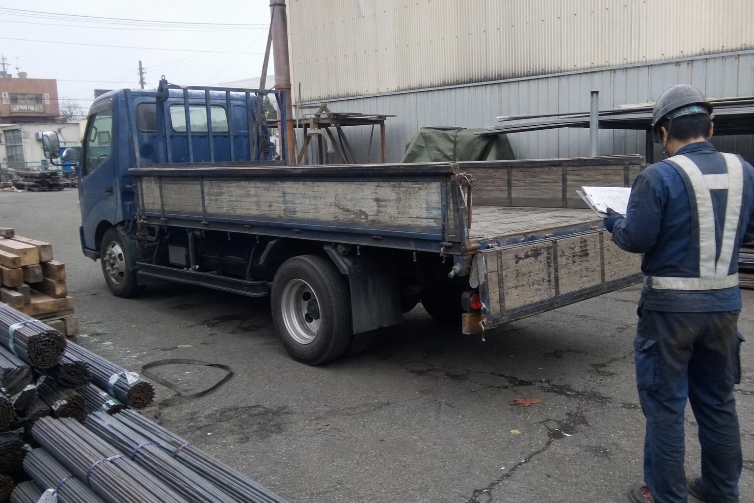 荷主の担当者が出荷伝票を確認しながら、鋼材と長尺物を積む平ボディトラックの荷台を点検している様子